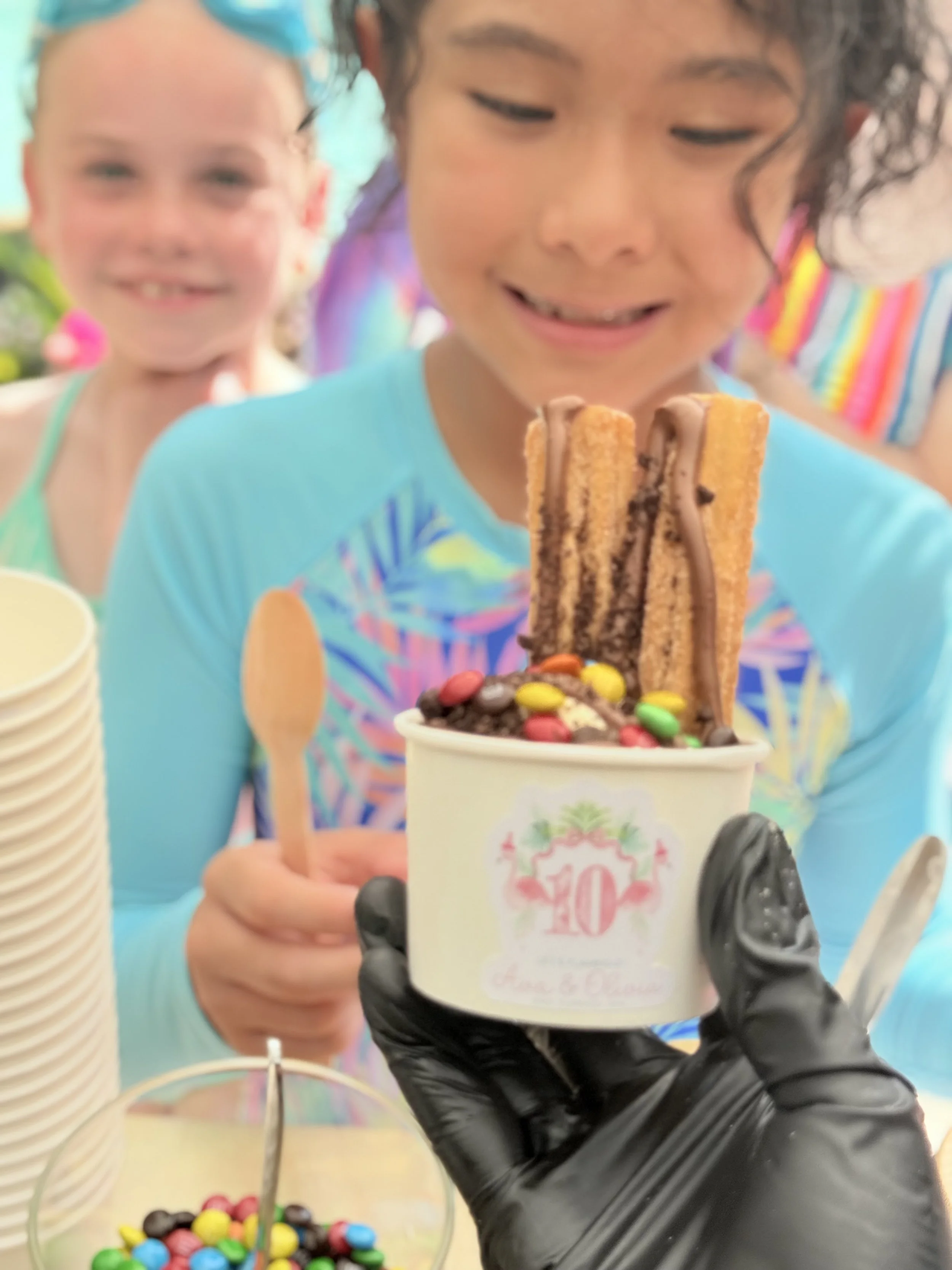 A young girl in a blue shirt receiving ice cream topped with Oreo cookies, mini M&Ms, and a wafer stick, with other children and birthday decorations in the background.