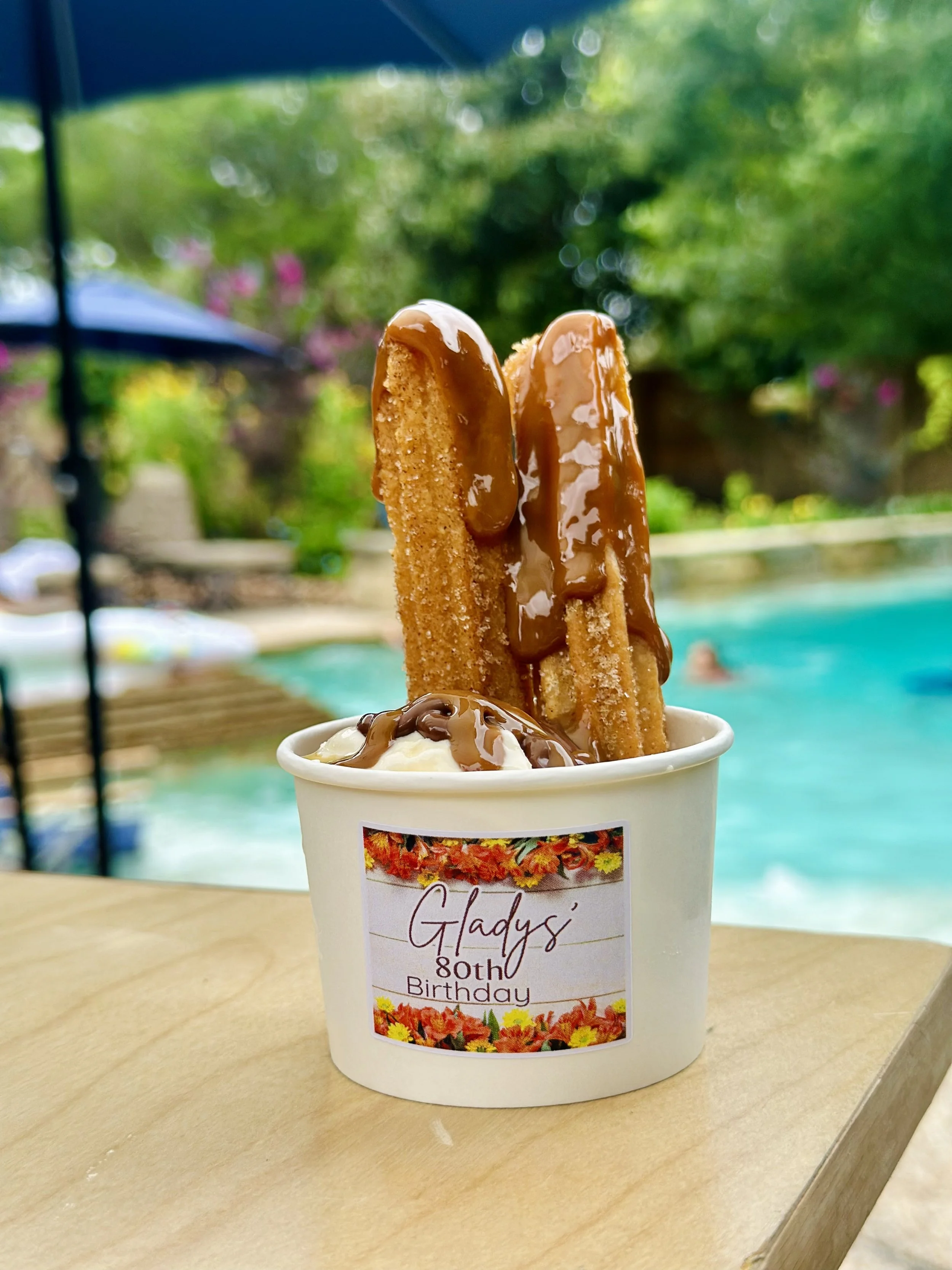 Ice cream topped with caramel sauce and two churros, placed in a cup with a customized label for Glady's 80th birthday, next to a swimming pool with green trees and a pink flowering bush in the background.