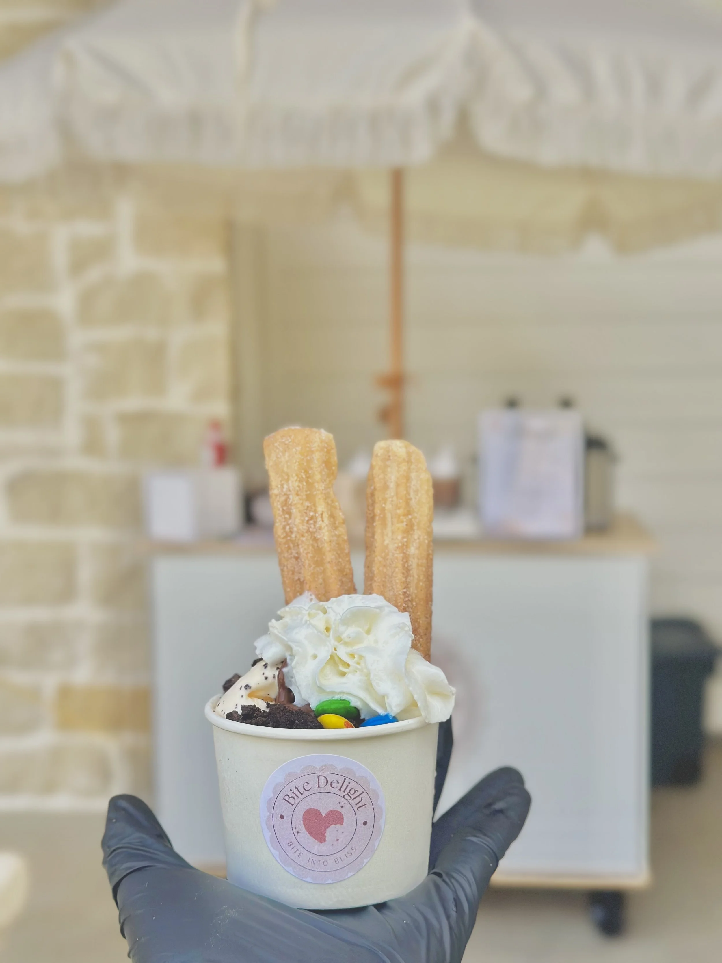 Cup of ice cream topped with whipped cream, two churros, rainbow-colored sprinkles, and chocolate cookie crumbs, held by a person wearing a black glove, indoors with a blurred background.