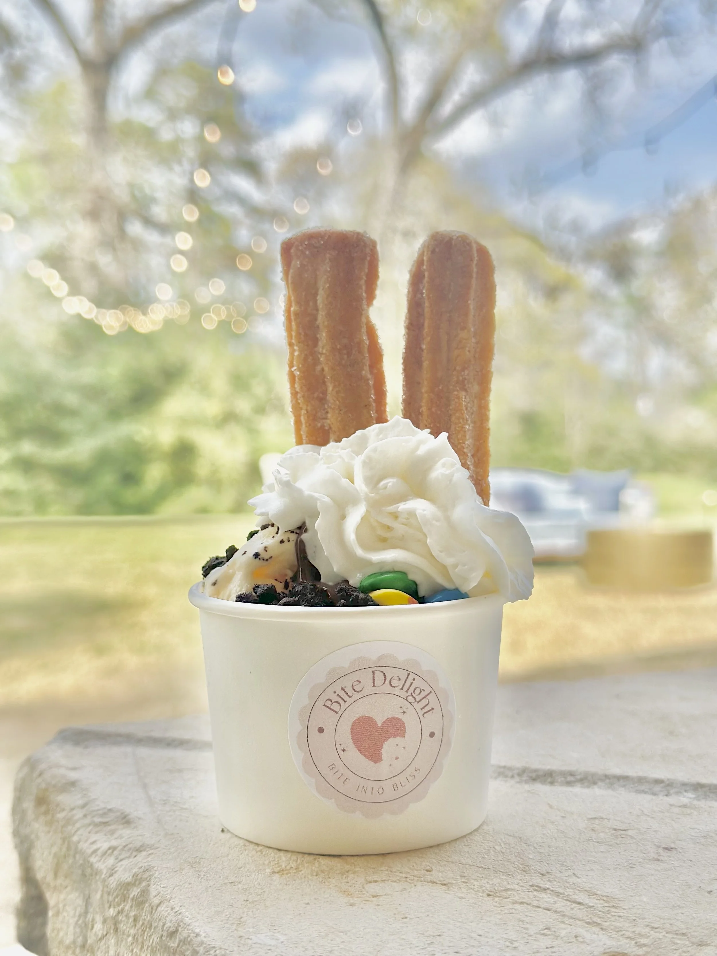 Cup of ice cream topped with whipped cream, chocolate cookie crumbles, colorful candy, and two churros, outside on a sunny day.