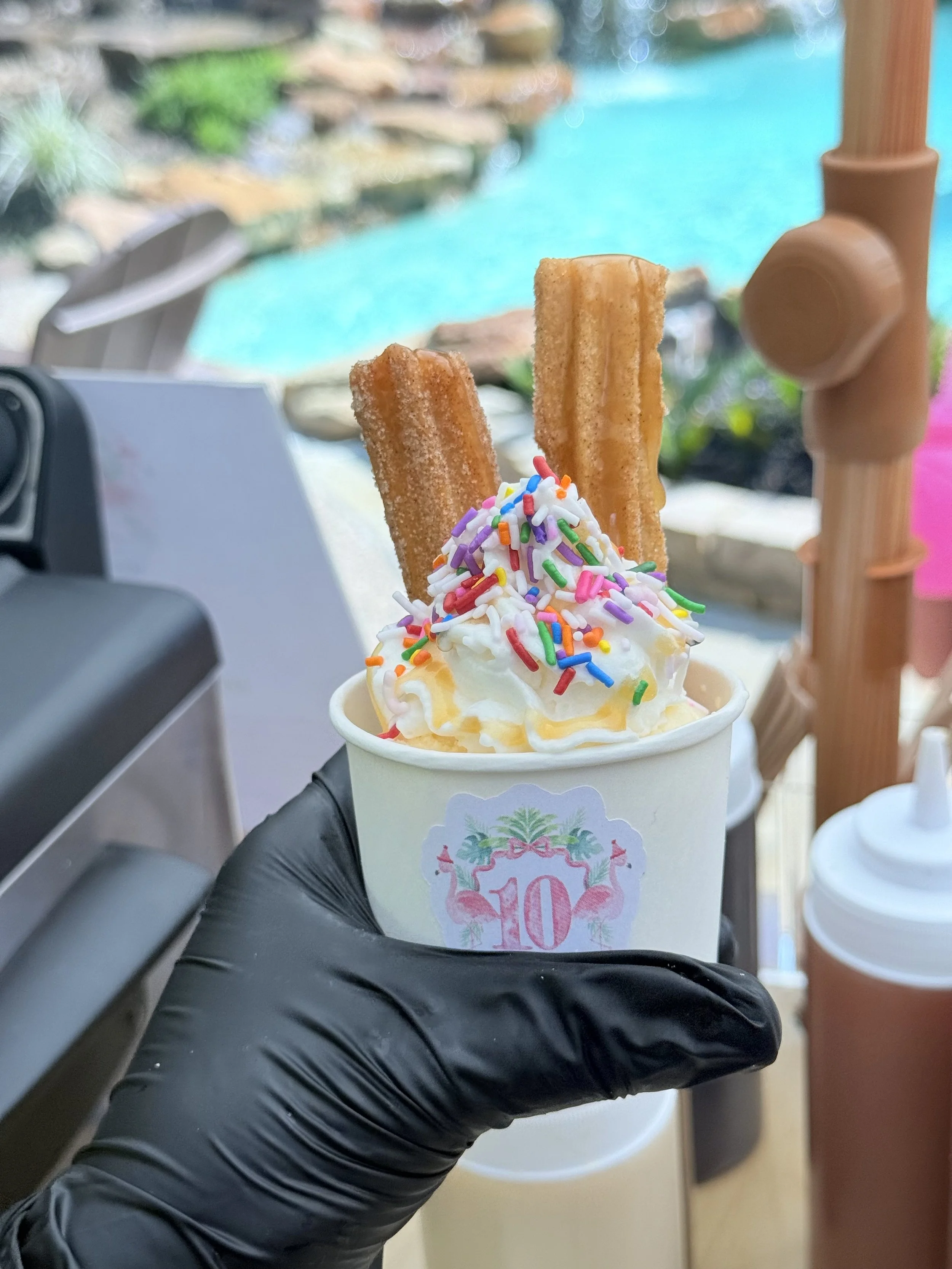 A cup of soft serve ice cream topped with colorful sprinkles, with two churros sticking out, held by a hand in a black glove.