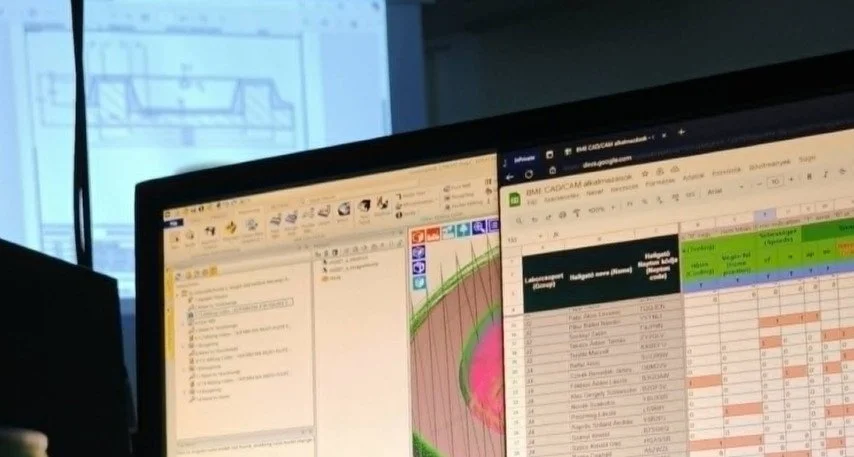 Two computer monitors displaying spreadsheets and data analysis, with a blurred background showing a window and part of a building exterior.