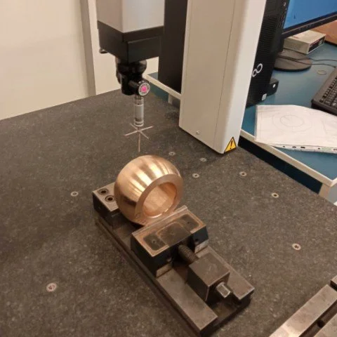 A precision measurement device with a metallic spherical object secured in a mechanical clamp, in a laboratory or workshop setting.