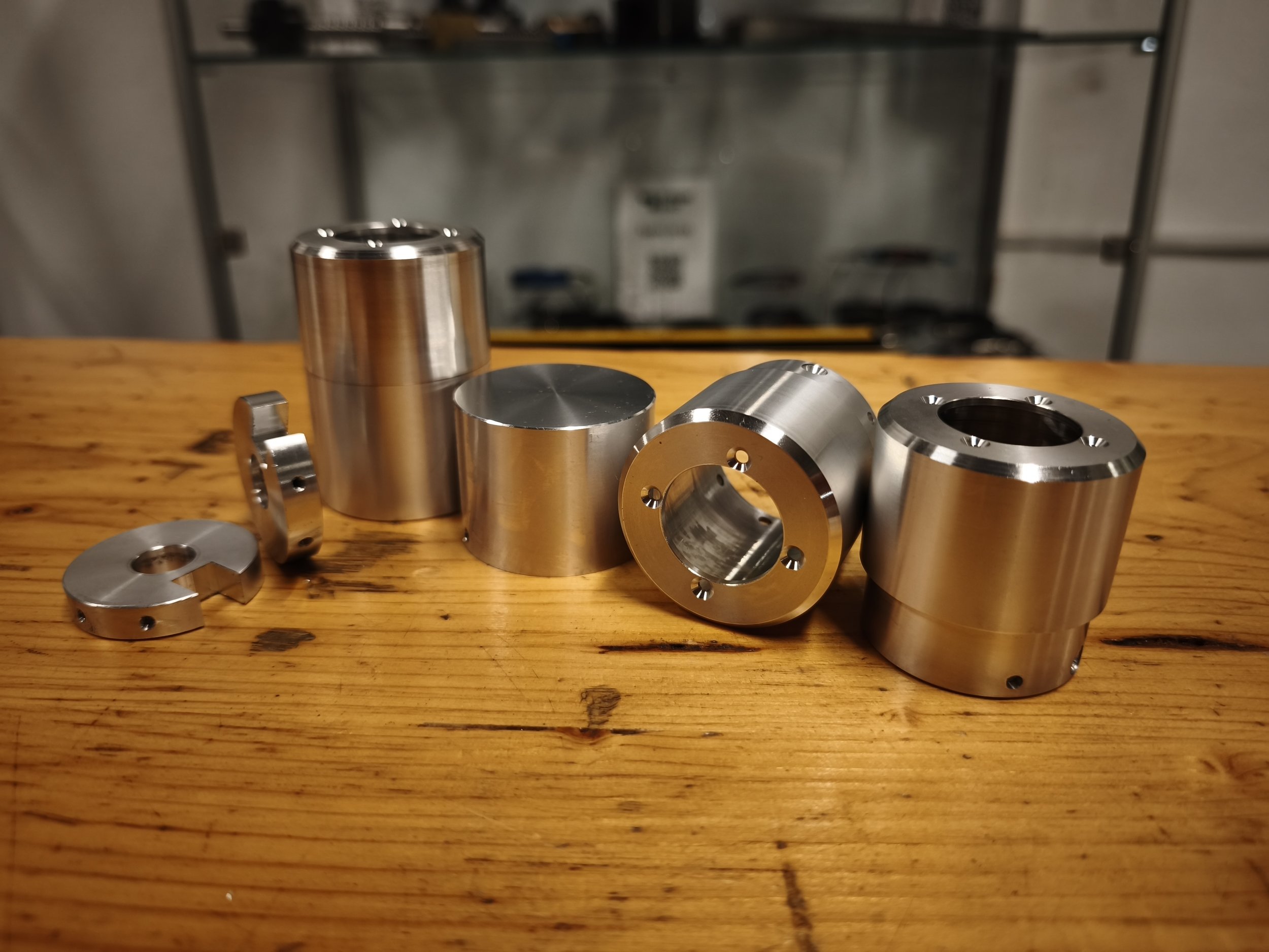 Several machined metal cylindrical components with holes and grooves on a wooden surface, with a blurred background of a shelving unit.