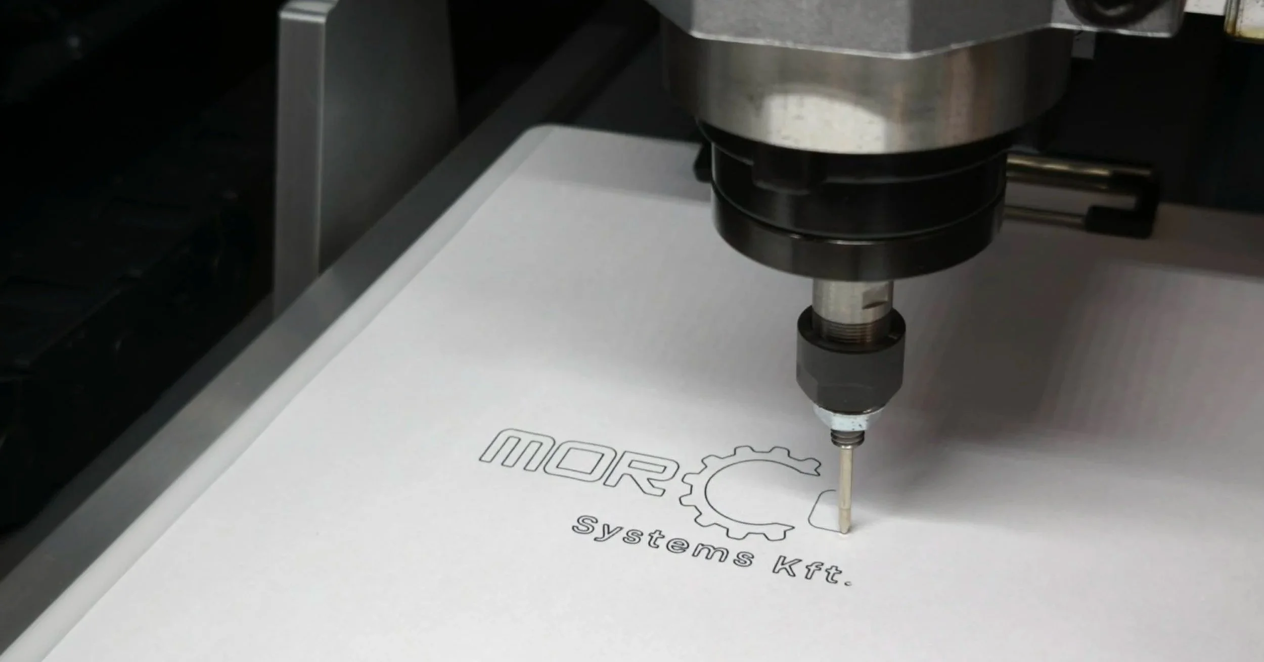 Close-up of a CNC machine engraving text and a gear logo on a white surface labeled 'MORA systems kft'.