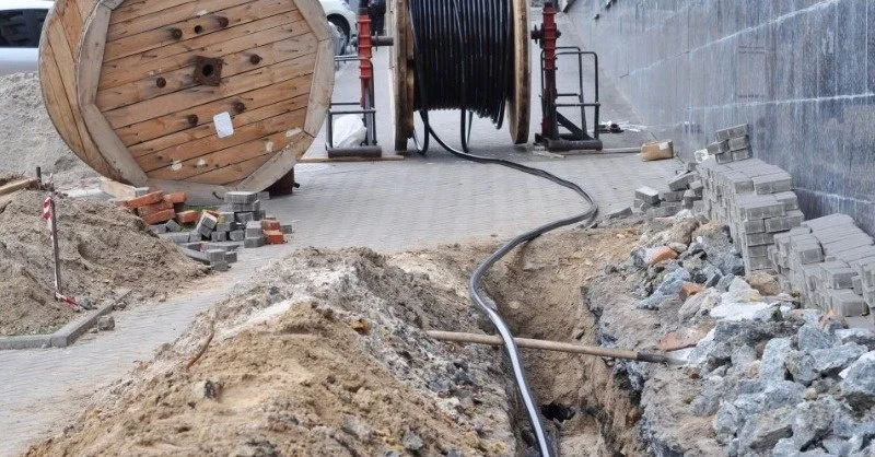 Construction site with a trench, piles of dirt and rocks, a large wooden spool of cable, and a reel of black conduit or cable in the background.
