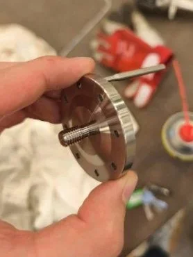 Close-up of a person's hand holding a metal machined part with a threaded bolt, with workshop tools and materials in the background.