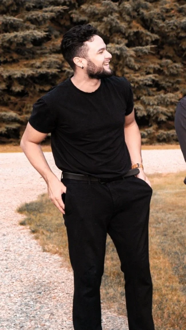 A young man with short dark hair, a beard, and earrings, smiling and wearing a black t-shirt and black pants, standing outdoors in front of pine trees.