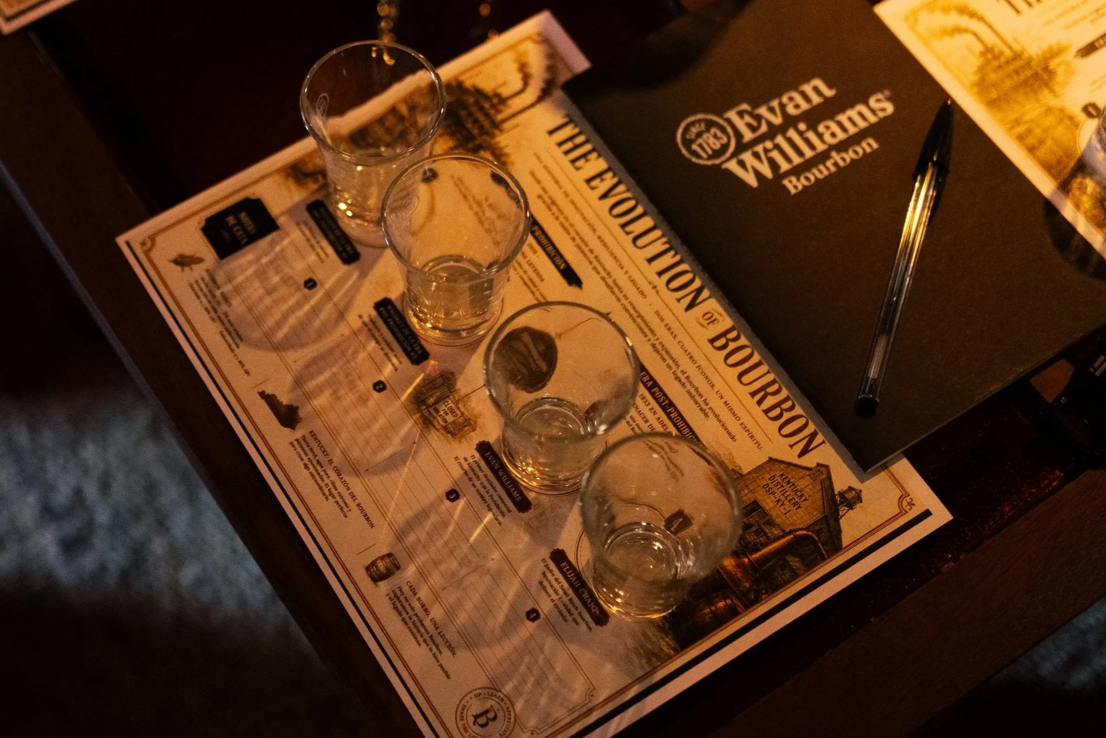 Mesa con varios vasos de diferentes tamaños y un folleto de eventos de bourbon en el centro, con un bolígrafo negro a un lado.