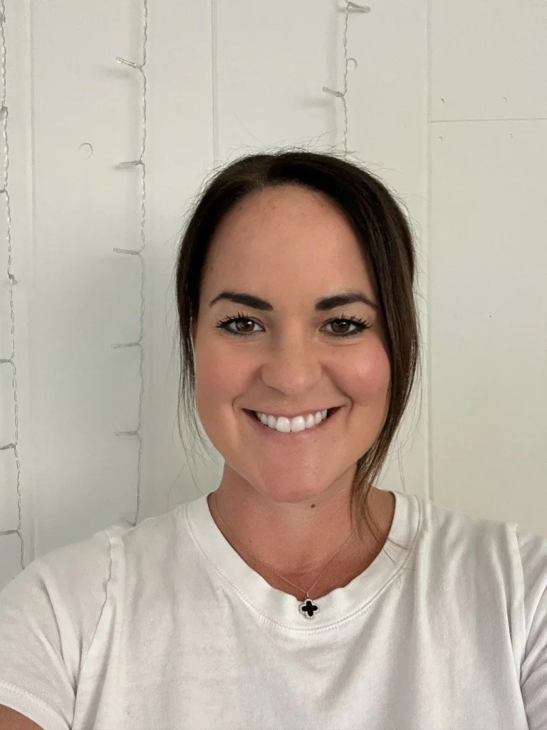 A woman with dark brown hair, fair skin, and a friendly smile taking a selfie indoors with a white wall and string lights in the background.