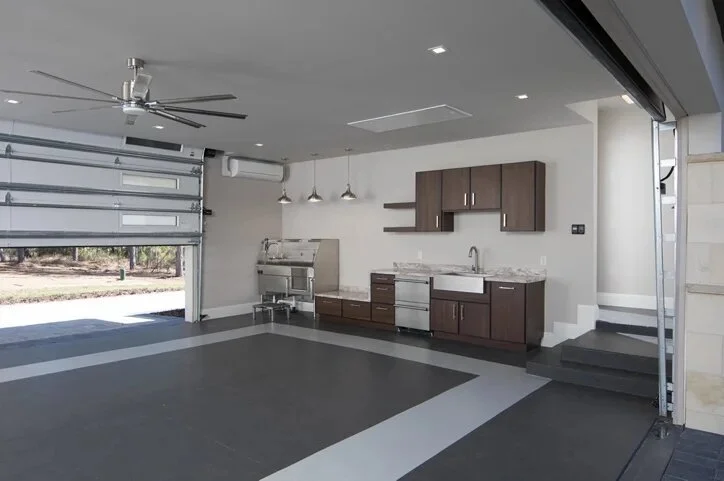 A well-organized modern garage with a kitchenette featuring stainless steel appliances and brown cabinets. The garage has an open door, a ceiling fan, overhead lighting, and steps leading to a raised platform area.