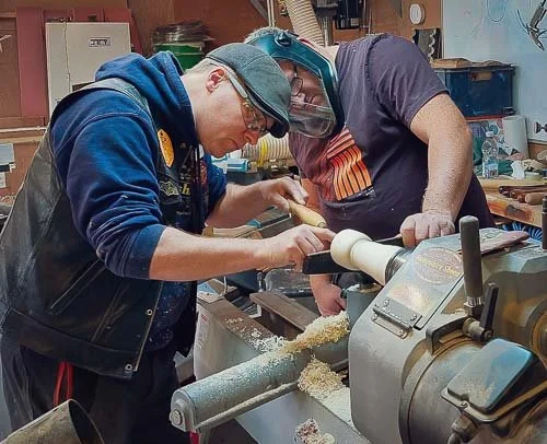Two men wearing protective helmets and glasses, working together on a woodworking project in a workshop. One man is guiding the other's hands on a lathe machine.