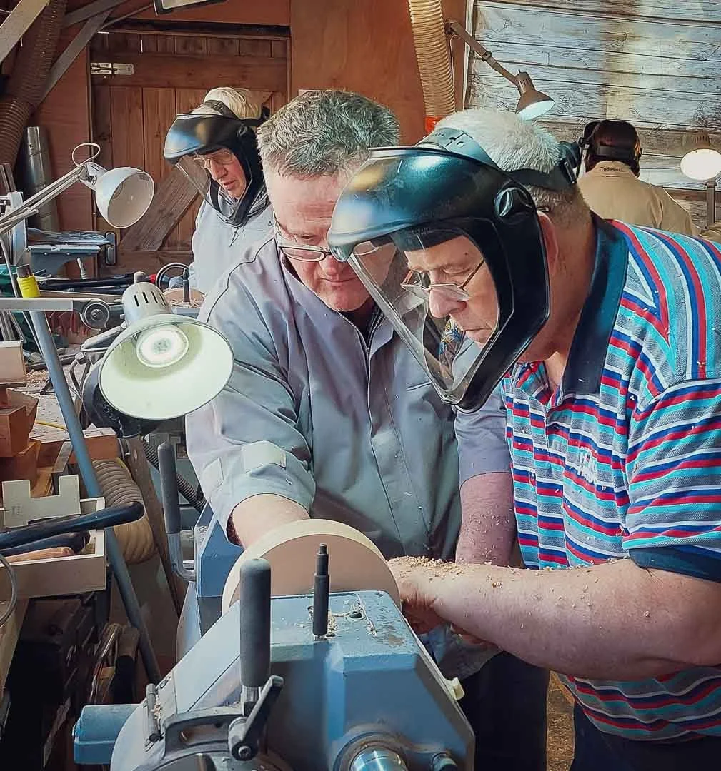 Three men wearing safety helmets working together on a woodworking lathe in a workshop, with various lamps and tools around them.