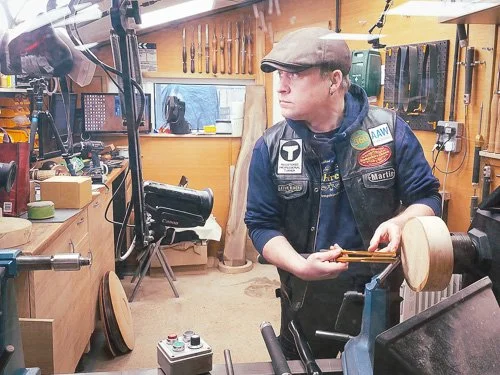 A man working in a woodworking shop, using a lathe to shape a piece of wood, with woodworking tools and equipment in the background.