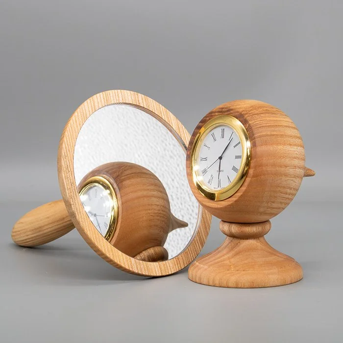 A wooden clock with a round face and Roman numerals, placed on a wooden stand, with a round mirror reflecting the clock and background.