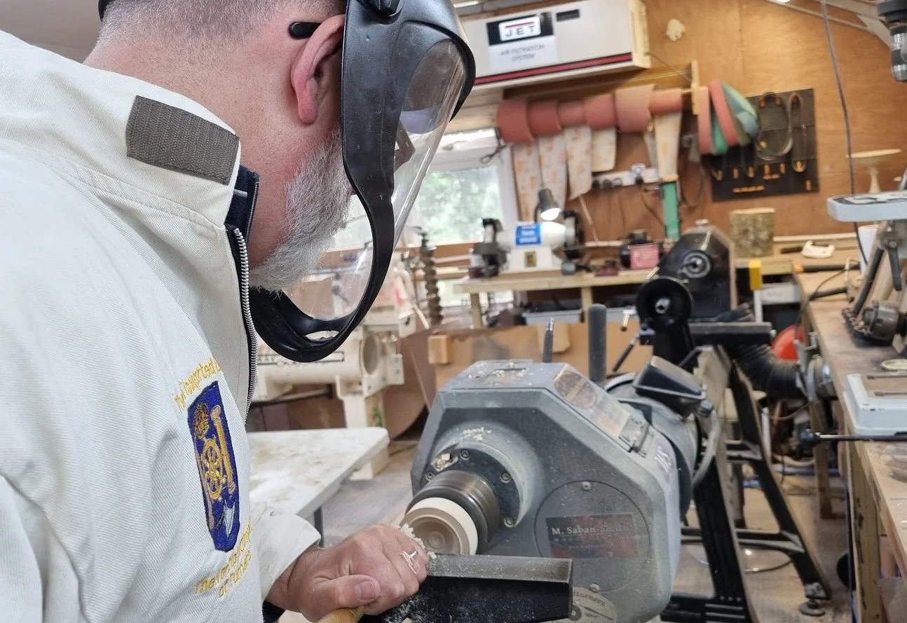 A man wearing safety goggles and a white shirt working with a wood lathe in a woodworking workshop, surrounded by tools and equipment.
