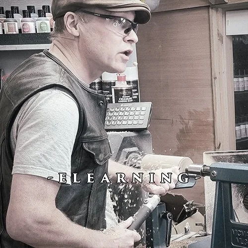 A man working in a woodworking shop, using a lathe to shape a piece of wood, with woodworking tools and equipment in the background.