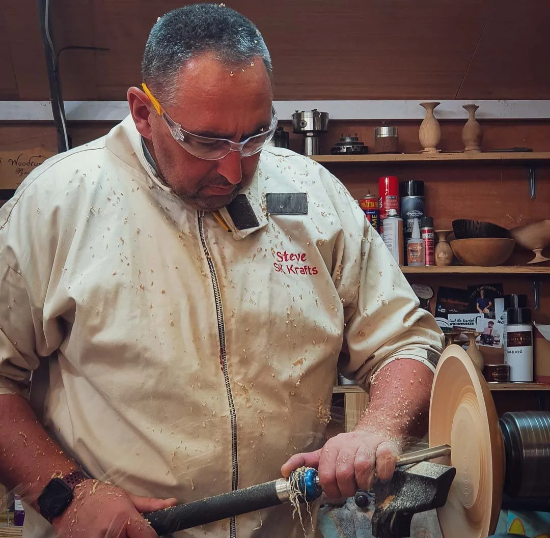 A man wearing safety glasses and a beige woodworking shirt engraved with 'Steve SK Krafts' is focusing on shaping a wooden bowl on a lathe. The workshop has shelves with various woodworking tools and supplies in the background.