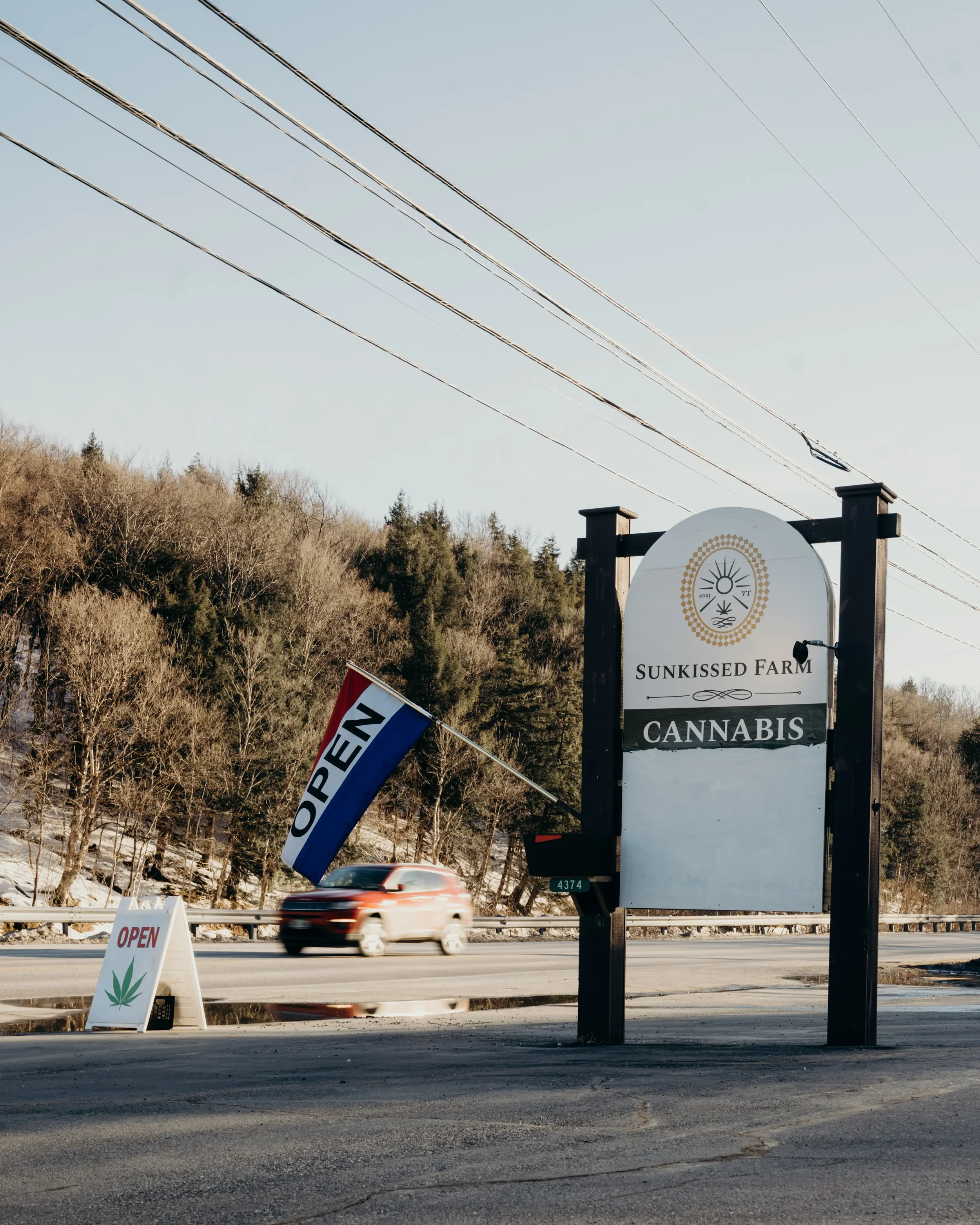 Sign for Sunkissed Farm Cannabis with an 'Open' flag and a smaller 'Open' sign with a cannabis leaf symbol at the roadside, with blurred car passing on the highway and trees in the background.
