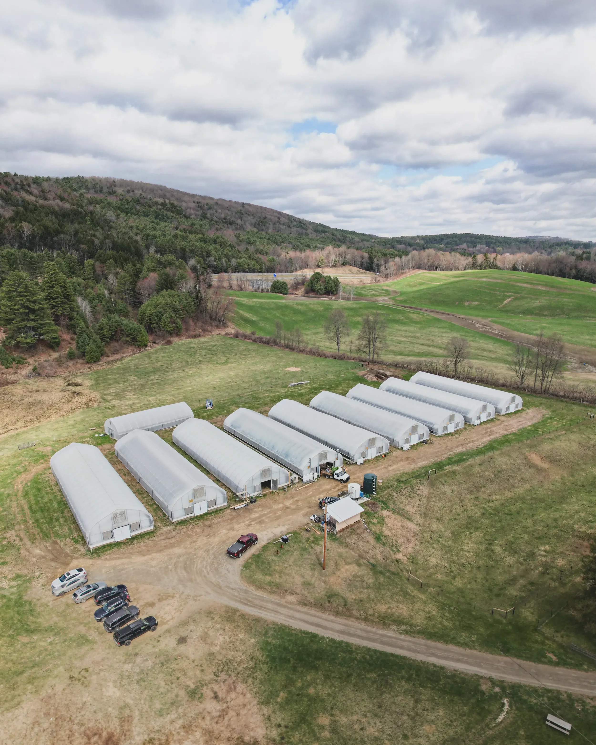 Aerial view of greenhouses, parked cars, and rolling hills with trees and fields in the background.