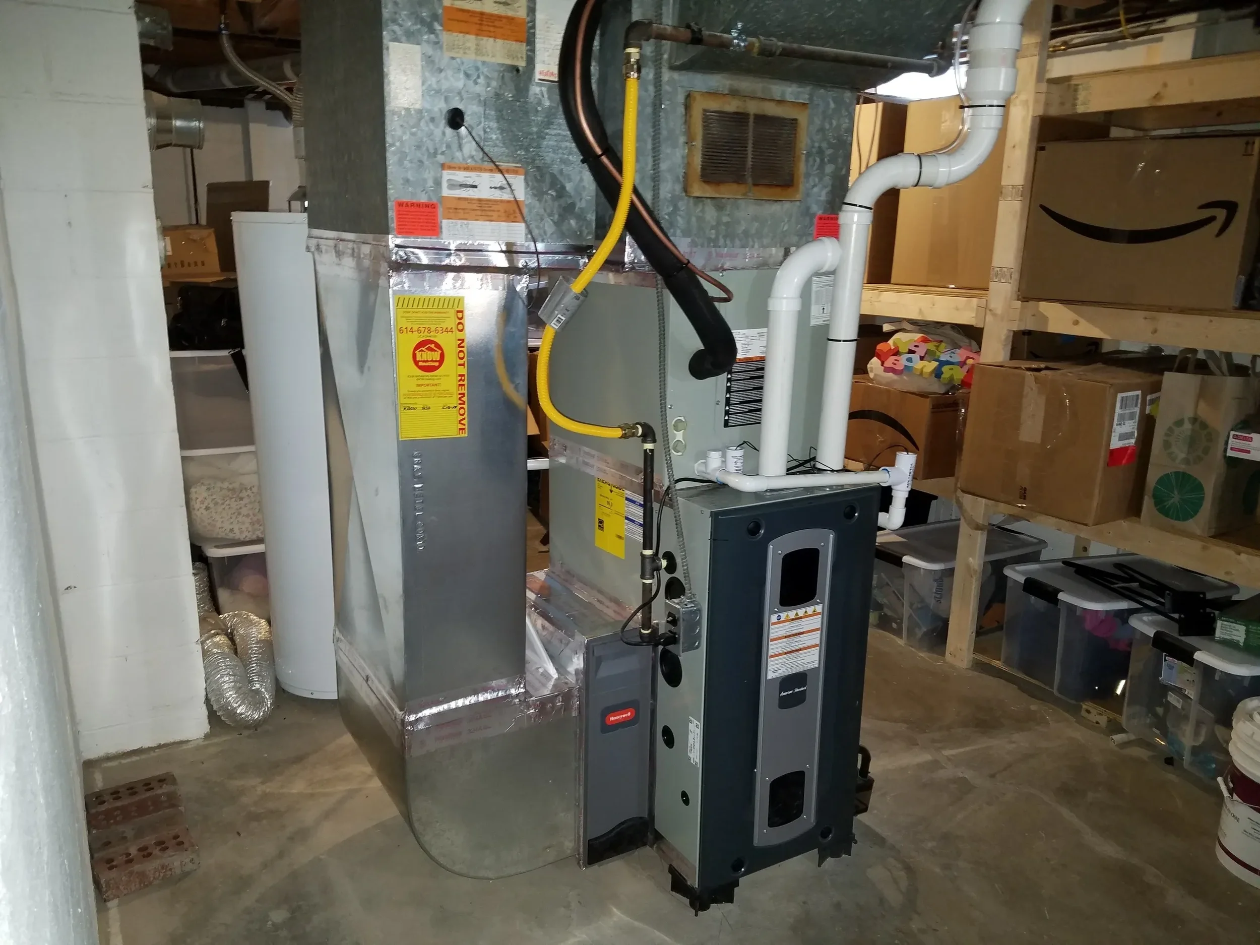 HVAC furnace system with ductwork and piping in a basement utility room, with shelves and storage boxes in the background.