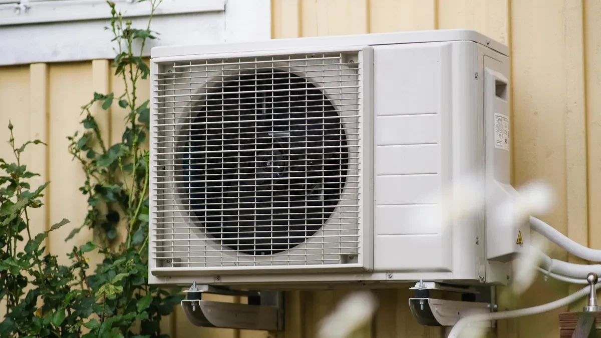 Outdoor air conditioning unit mounted on a wall with beige siding, surrounded by green plants.