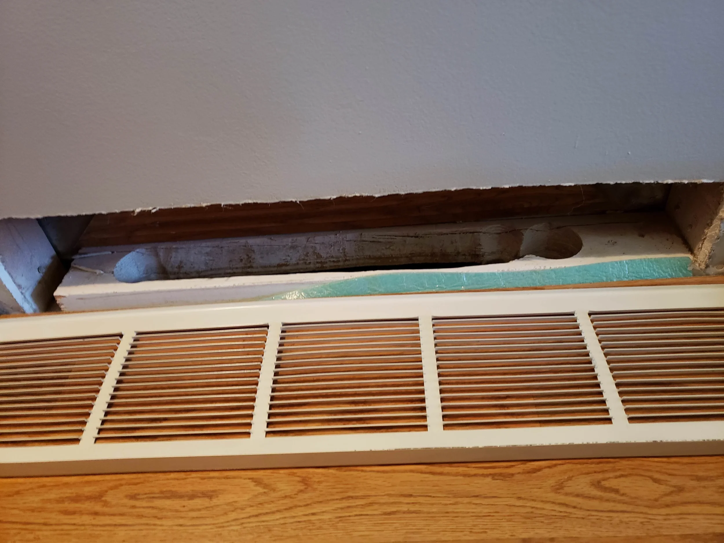 Close-up of a wall cleat and the bottom of a door with a vent cover. The vent cover is white with horizontal slats, and the wall above shows a missing piece revealing the rough interior wall with some wood framing and insulation.