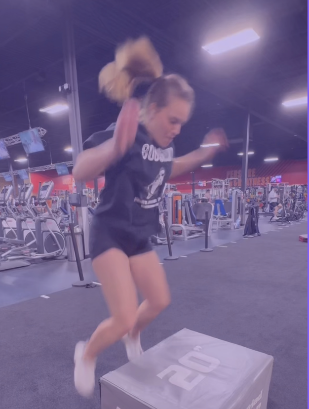 A young woman jumps over a 20-inch plyometric box during a workout at a gym, with exercise machines in the background.