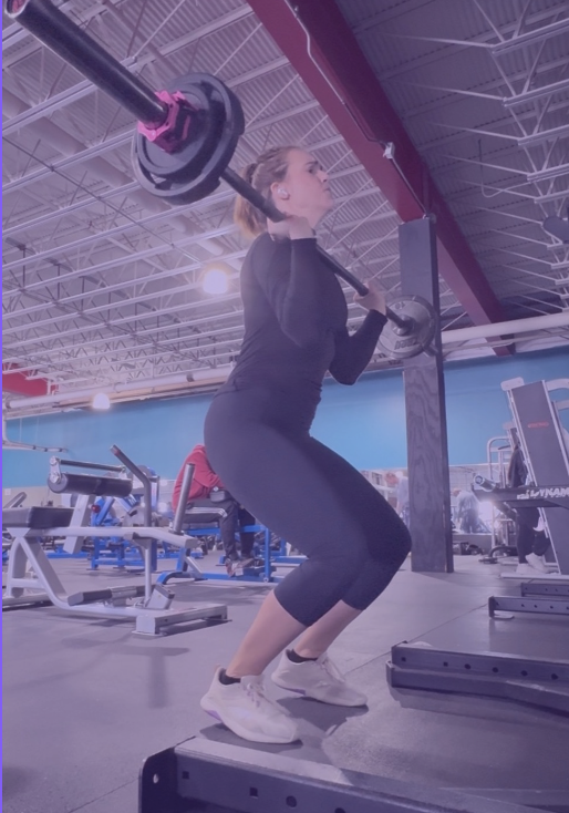 A woman lifting a barbell in a gym, standing on a platform, with workout equipment and other gym-goers in the background.