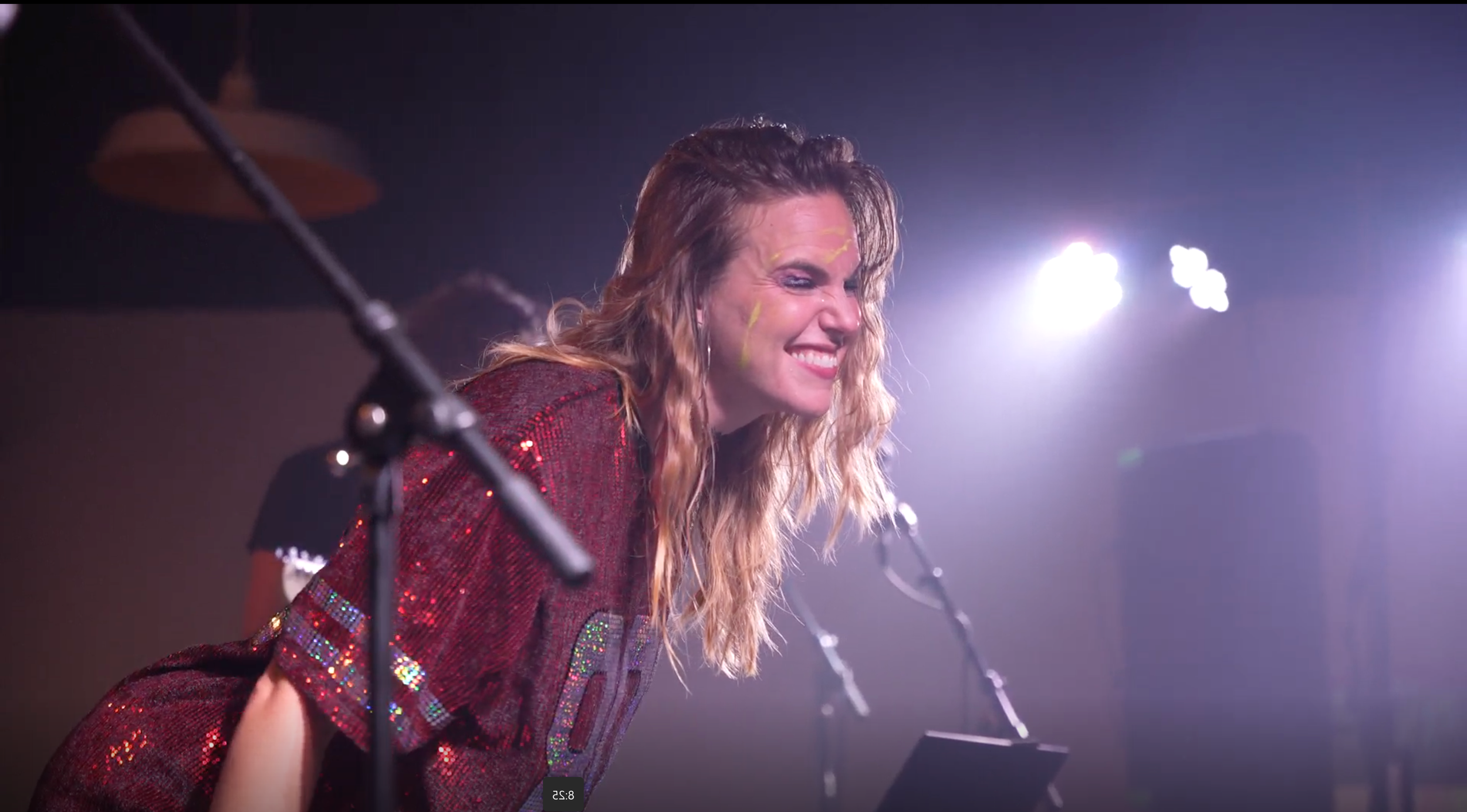 A woman with wavy blonde hair, wearing a sparkly red shirt, smiling and leaning forward on a stage with microphones in front of her, illuminated by bright stage lights.
