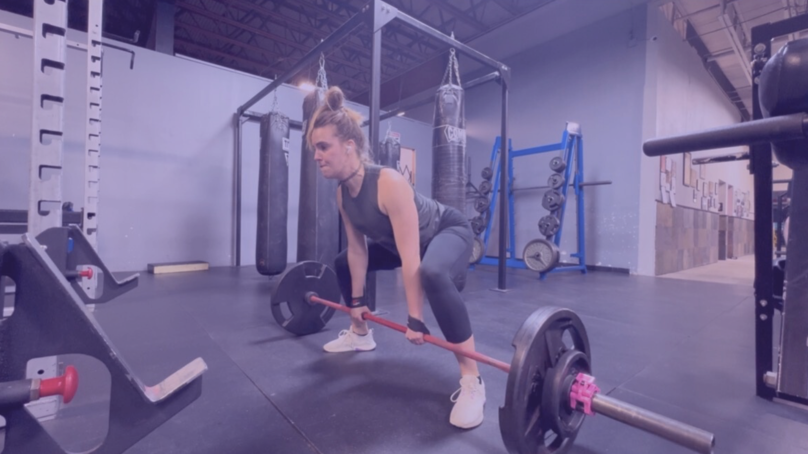 A woman in workout clothes lifting a barbell in a gym with punching bags and weight racks in the background.