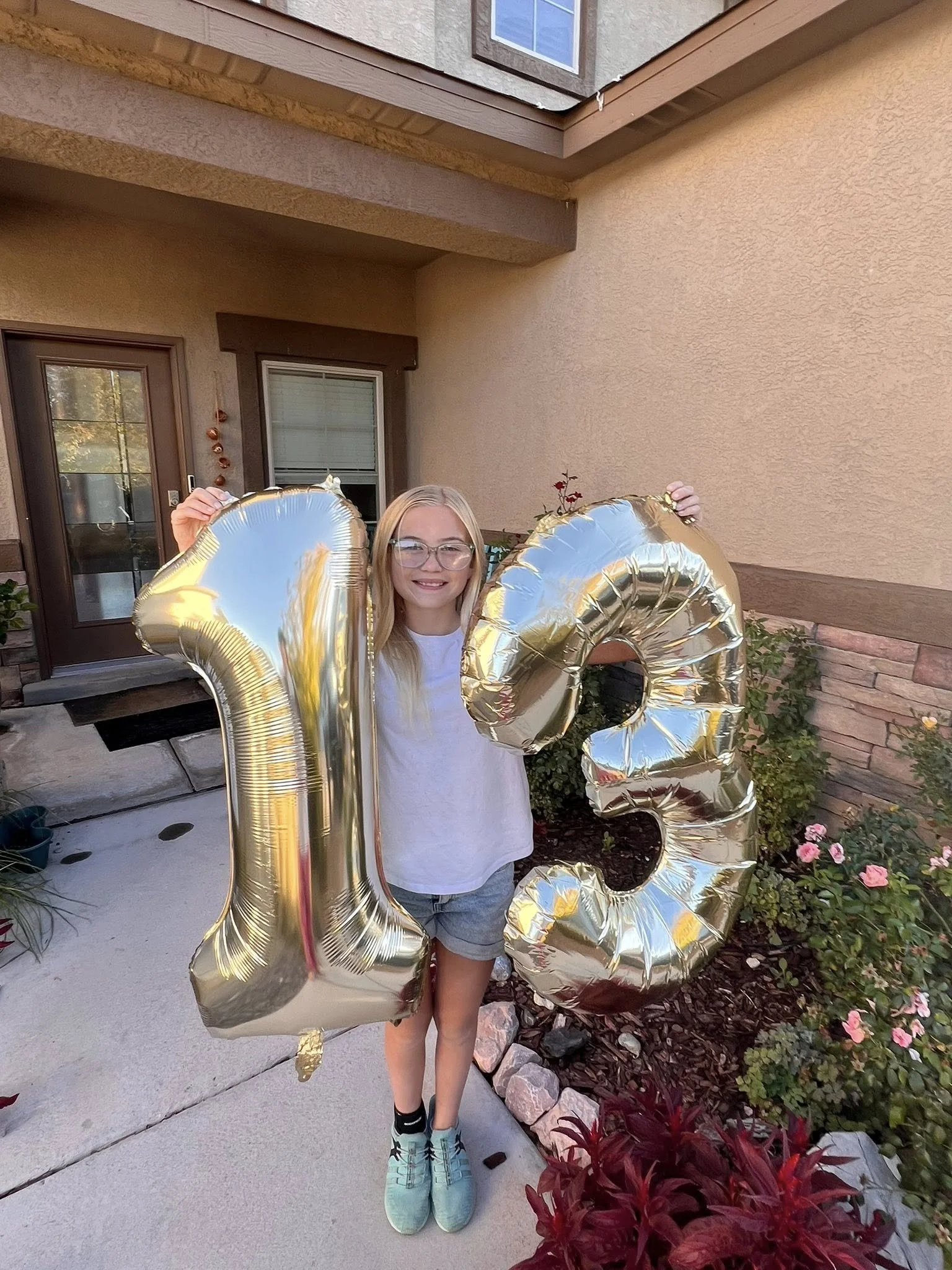 A young girl with blonde hair and glasses holding gold foil balloons in the shape of the numbers '1' and '3', standing outside in front of a house with a garden.