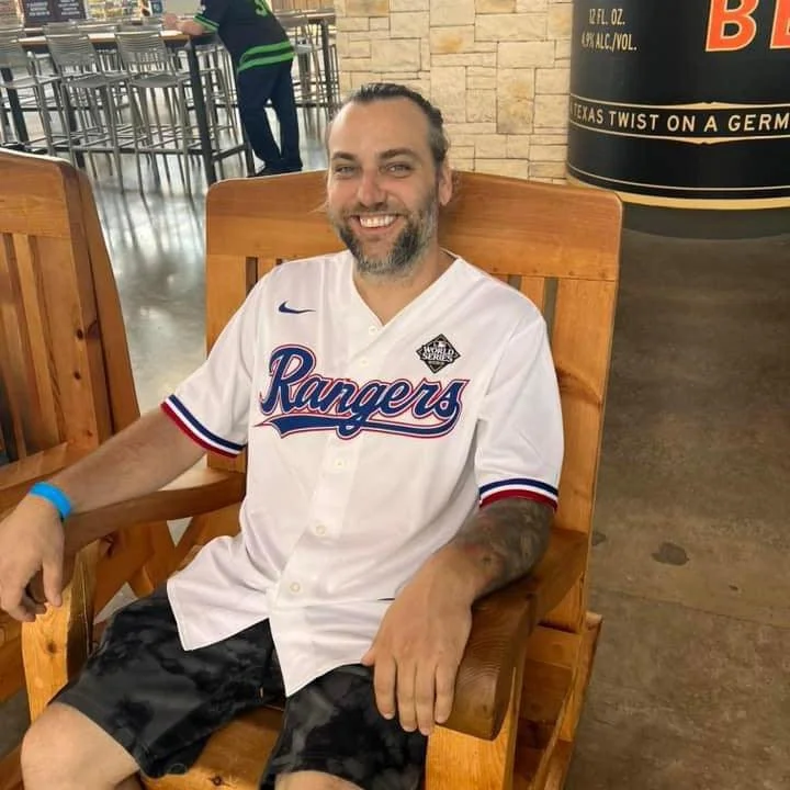 A man with a beard smiling and sitting on a wooden chair, wearing a white baseball jersey with 'Rangers' written on it and dark shorts, in a restaurant or bar setting.