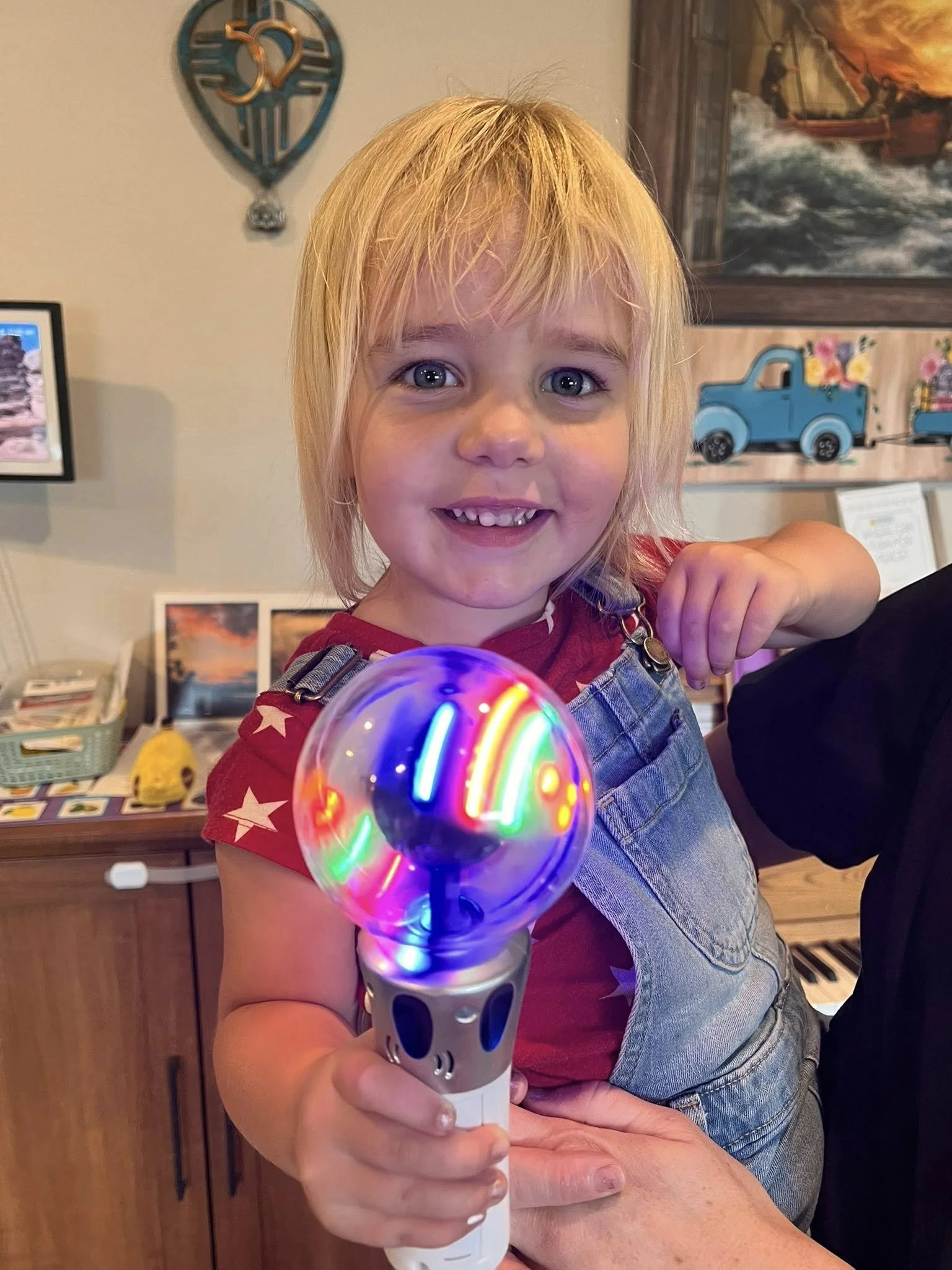 A young girl with blonde hair and blue eyes smiling while holding a colorful LED light wand in a living room.