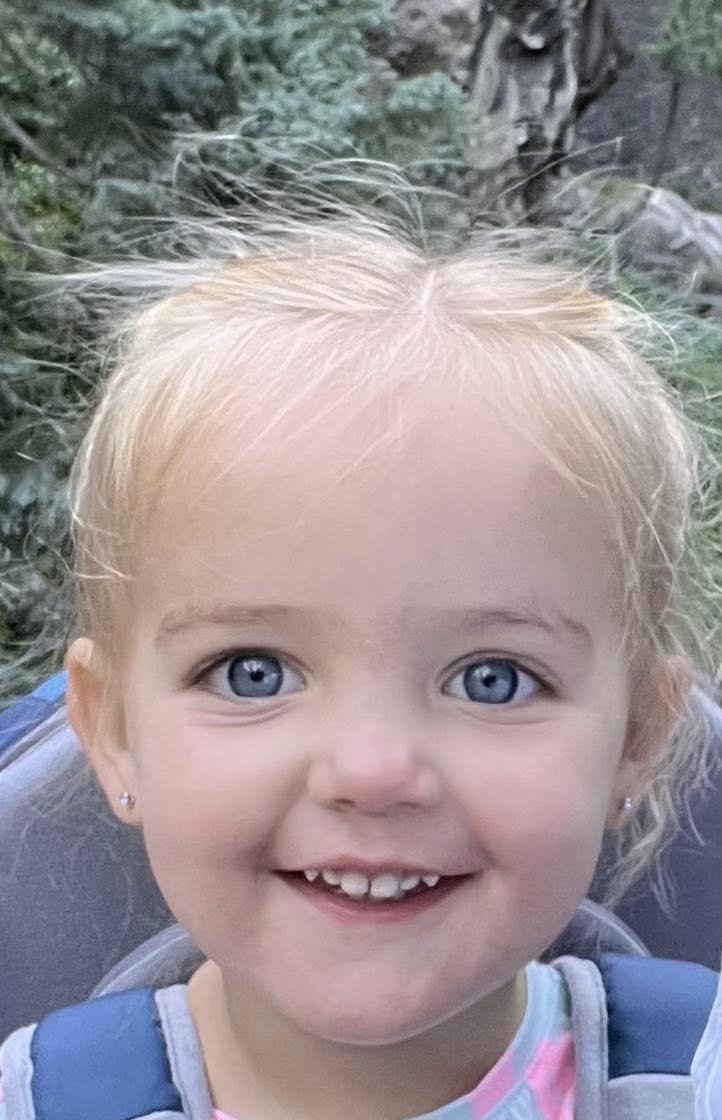 A young girl with blonde hair, blue eyes, and a big smile showing her teeth, outside surrounded by trees and greenery.