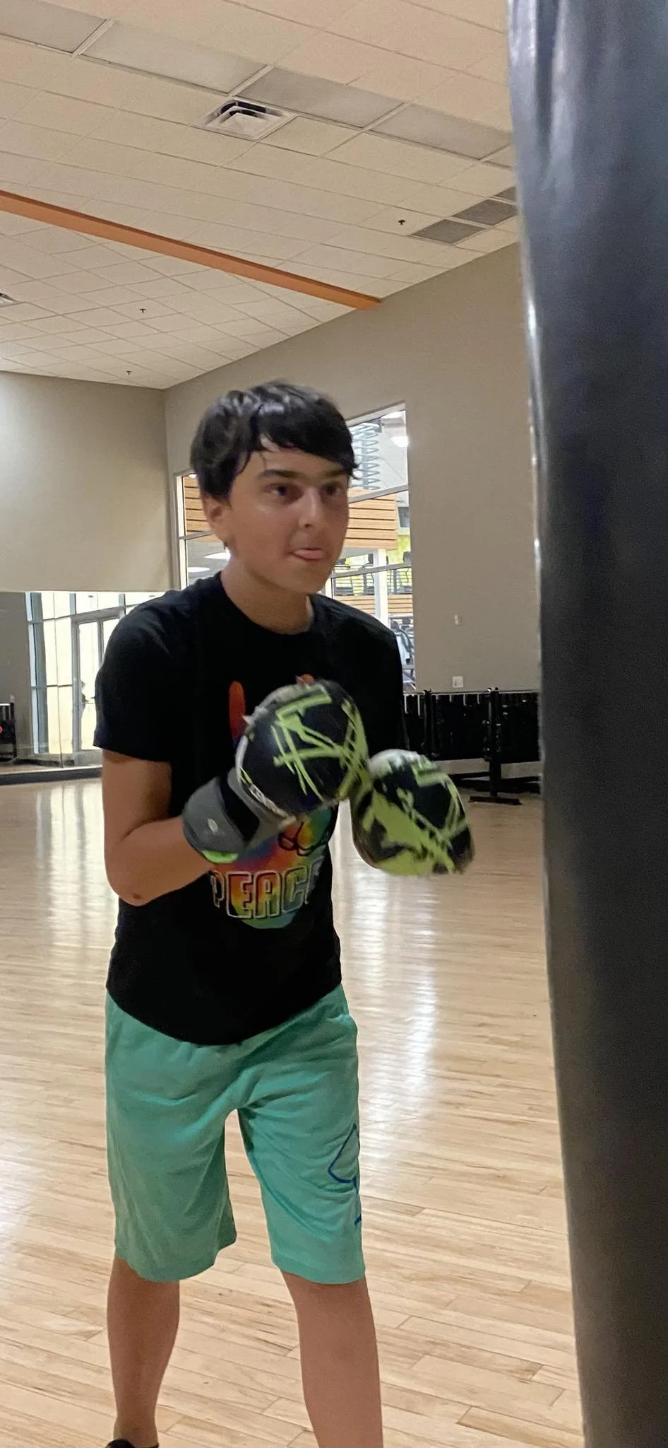 A young boy with black hair wearing a security guard t-shirt, green shorts, and boxing gloves in a gym with wooden flooring and large windows.