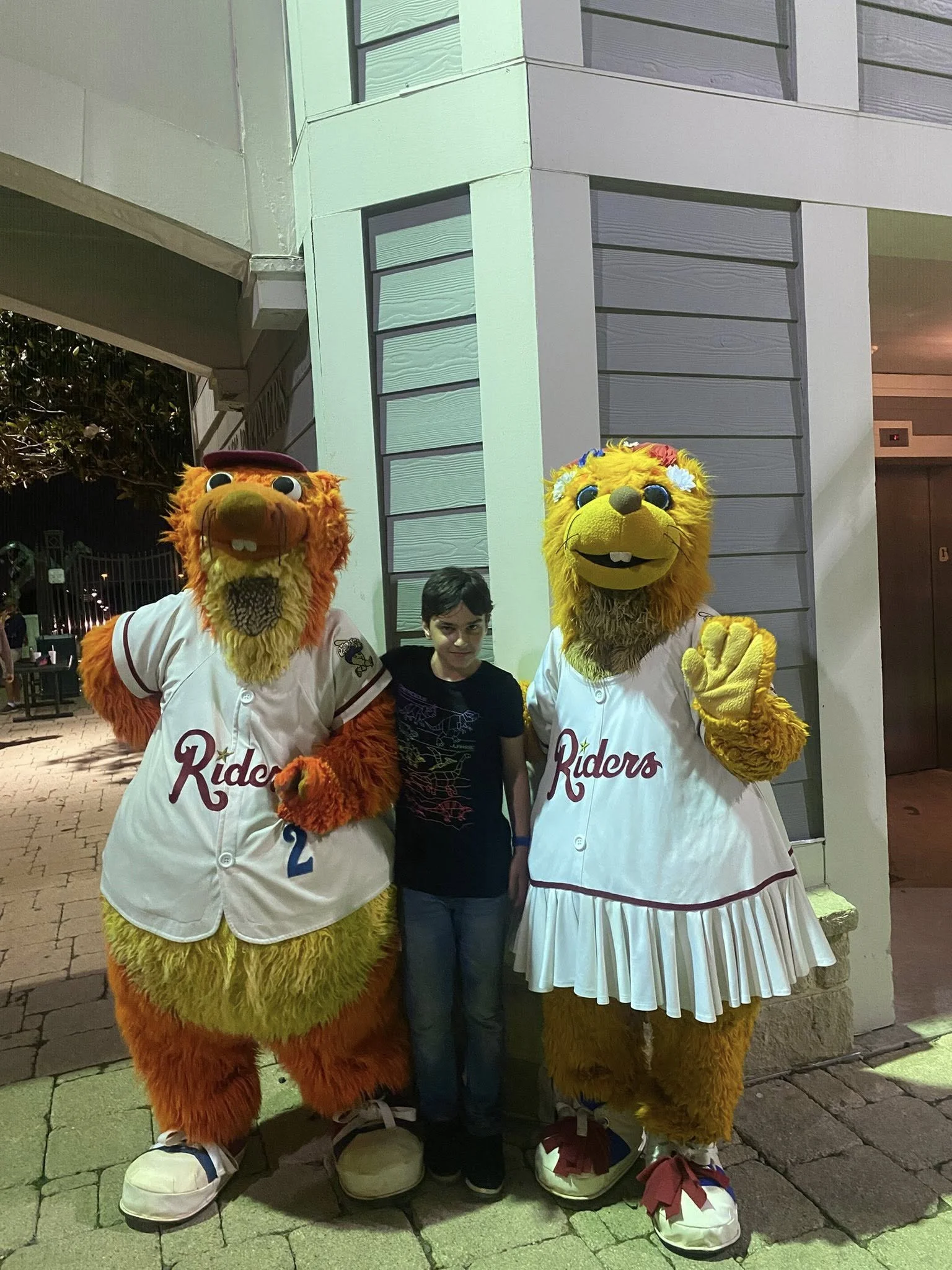 A young boy standing between two person in lion costumes dressed as baseball players, outside near a building at night.