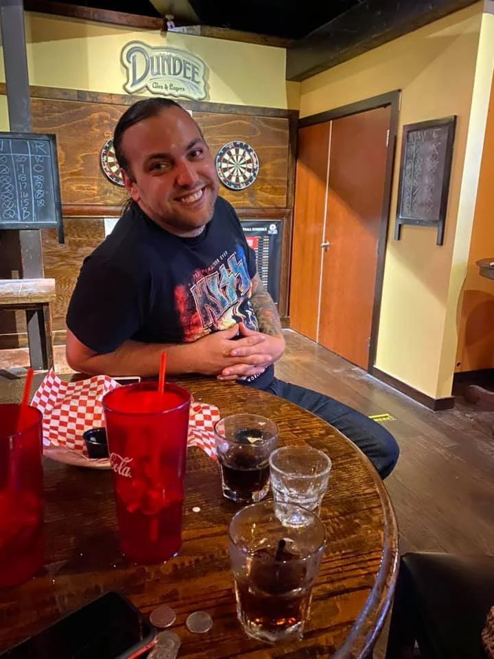 A smiling man with long dark hair, wearing a black Iron Maiden t-shirt, sitting at a wooden table in a bar or restaurant. The table has three glasses of soda, a few coins, and a black wallet. In the background, there's a dartboard, a menu on a chalkb