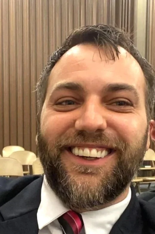Close-up of a smiling man with a beard, dressed in a suit and tie, in a conference or meeting room with wooden paneling and chairs in the background.