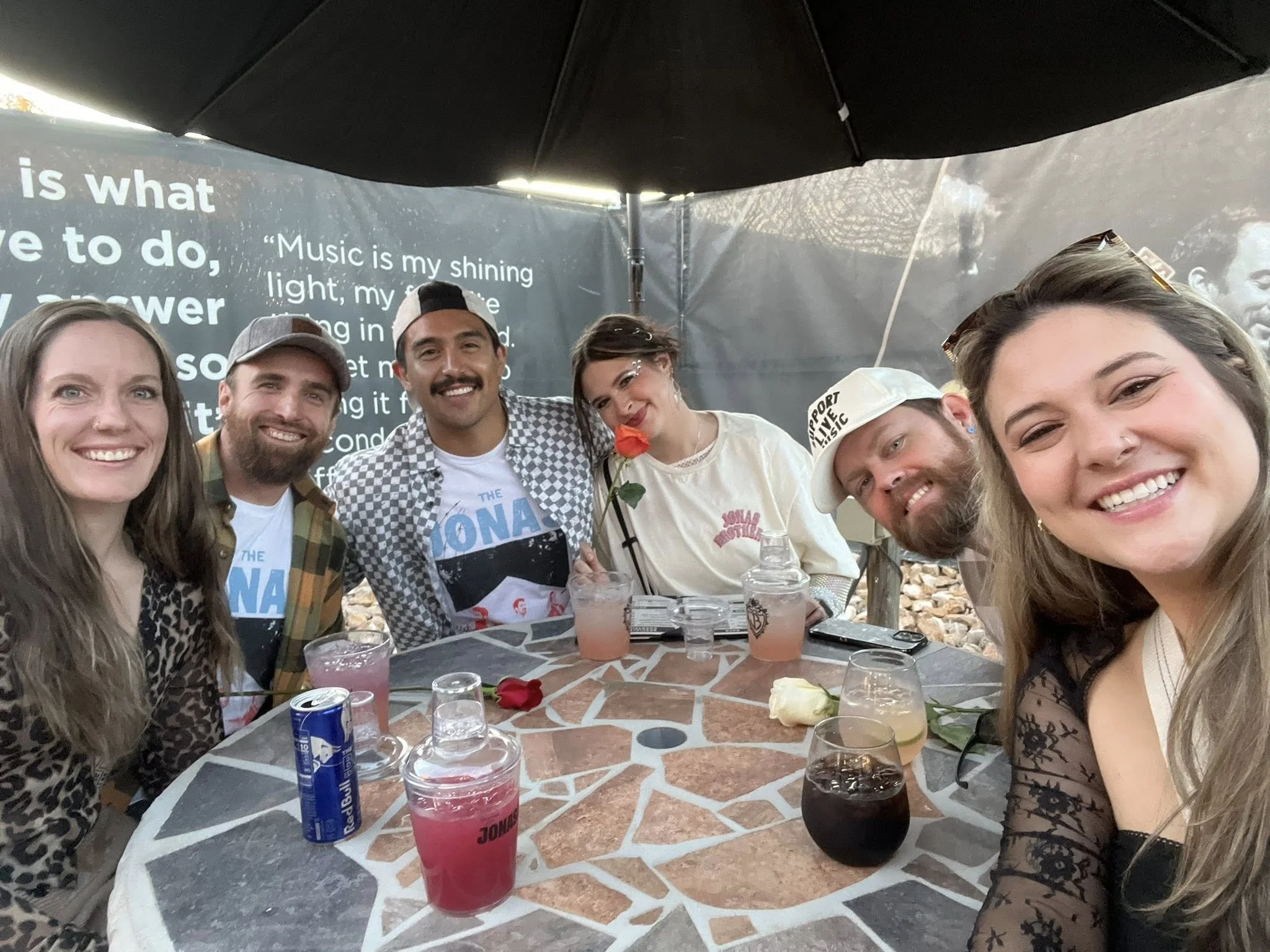 Group of six friends sitting around a table outdoors, smiling and enjoying drinks, with a black umbrella overhead and a banner with text in the background.