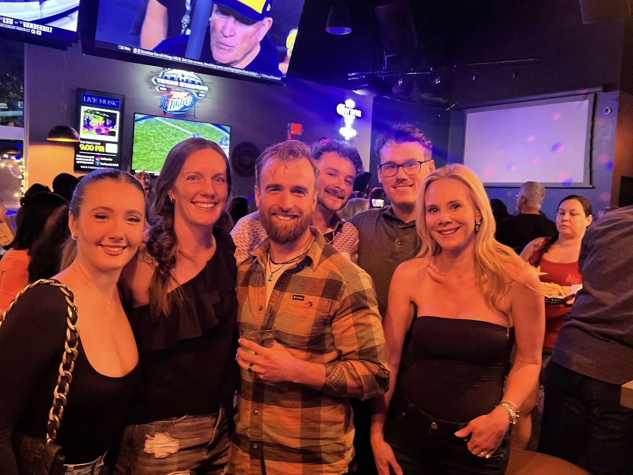 Group of six people posing together inside a lively bar or sports pub with television screens showing sports games in the background.