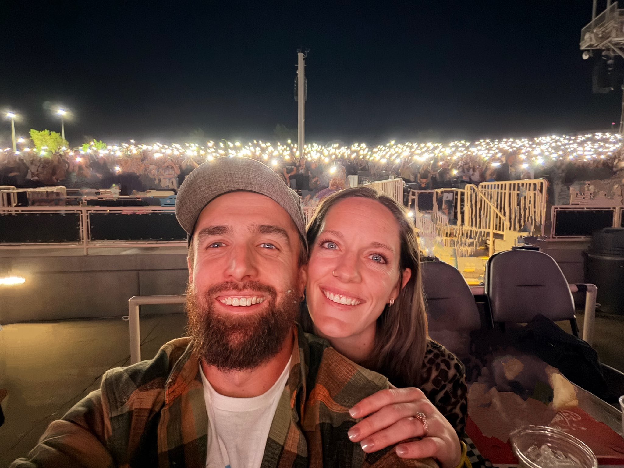 A smiling couple taking a selfie at night with an outdoor concert crowd illuminated by phone flashlights in the background.