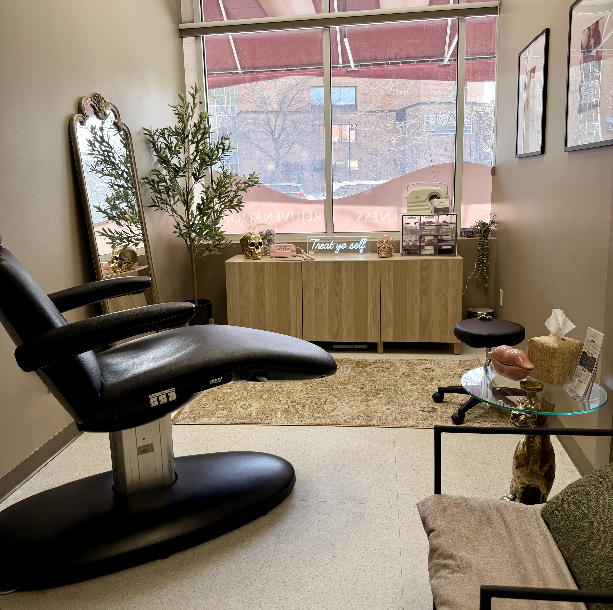 Interior of a beauty or skincare treatment room with a black treatment chair, a large mirror, a potted plant, and decorative skulls on a wooden cabinet. There is a neon sign that says "Treat yo self" and various aesthetic decor.