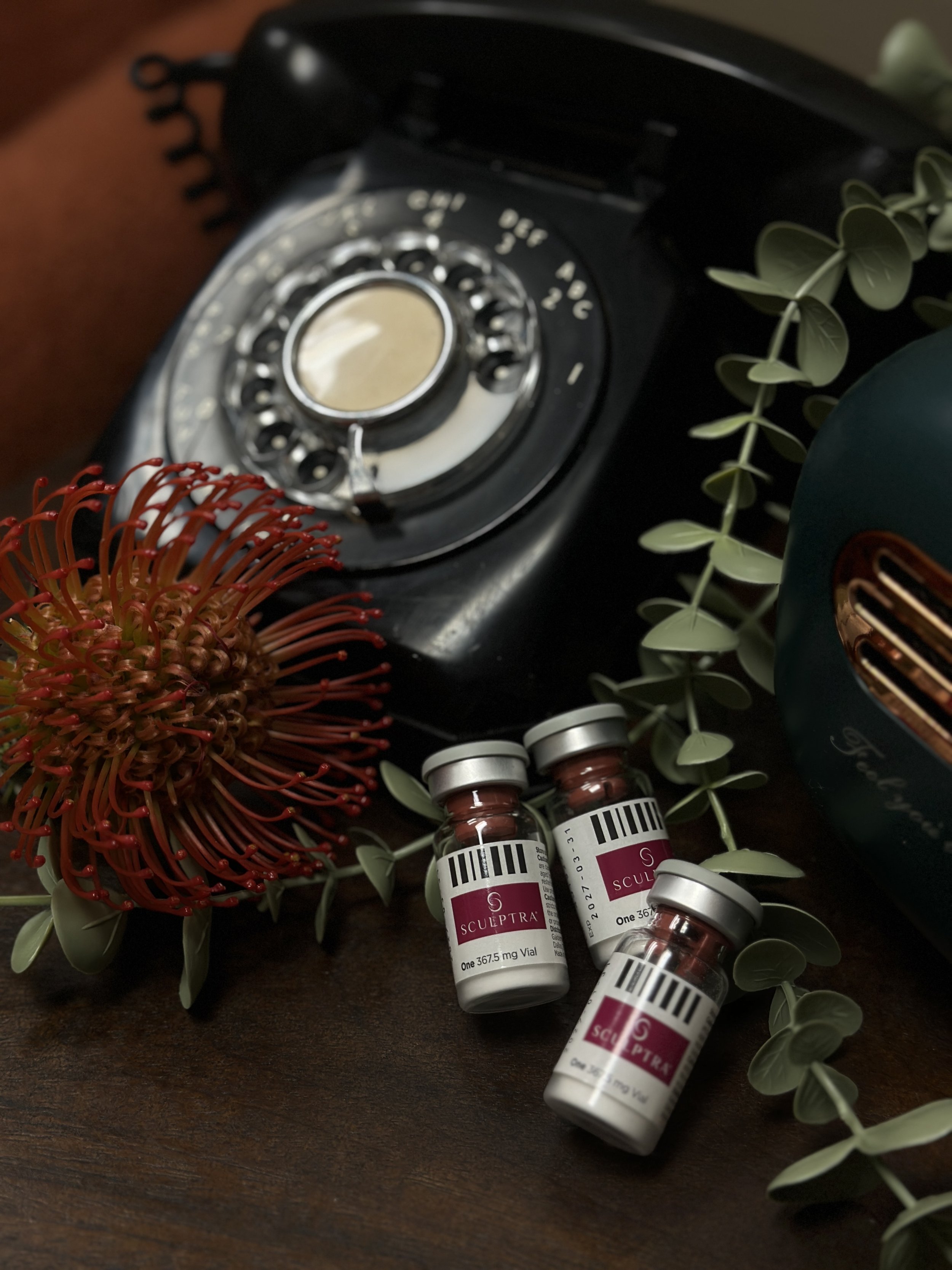 Vintage black rotary telephone surrounded by eucalyptus leaves, red flower, and three small vials of Sculptra injectable filler on a wooden surface.