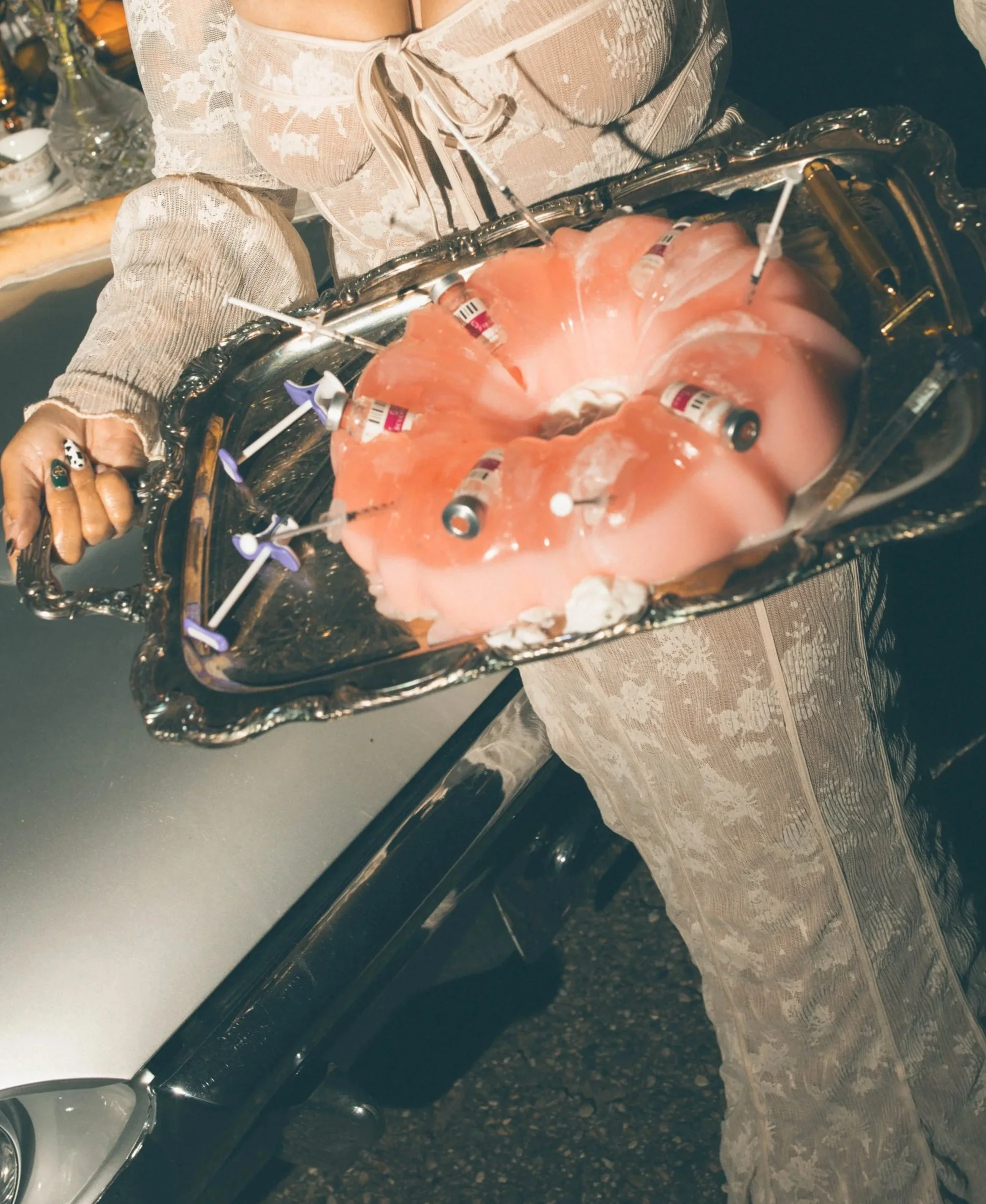 A person dressed in vintage lace clothing holding a silver tray with skull-shaped chamber filled with pink gelatin, syringes, and small vials, resembling a festive or theatrical display.