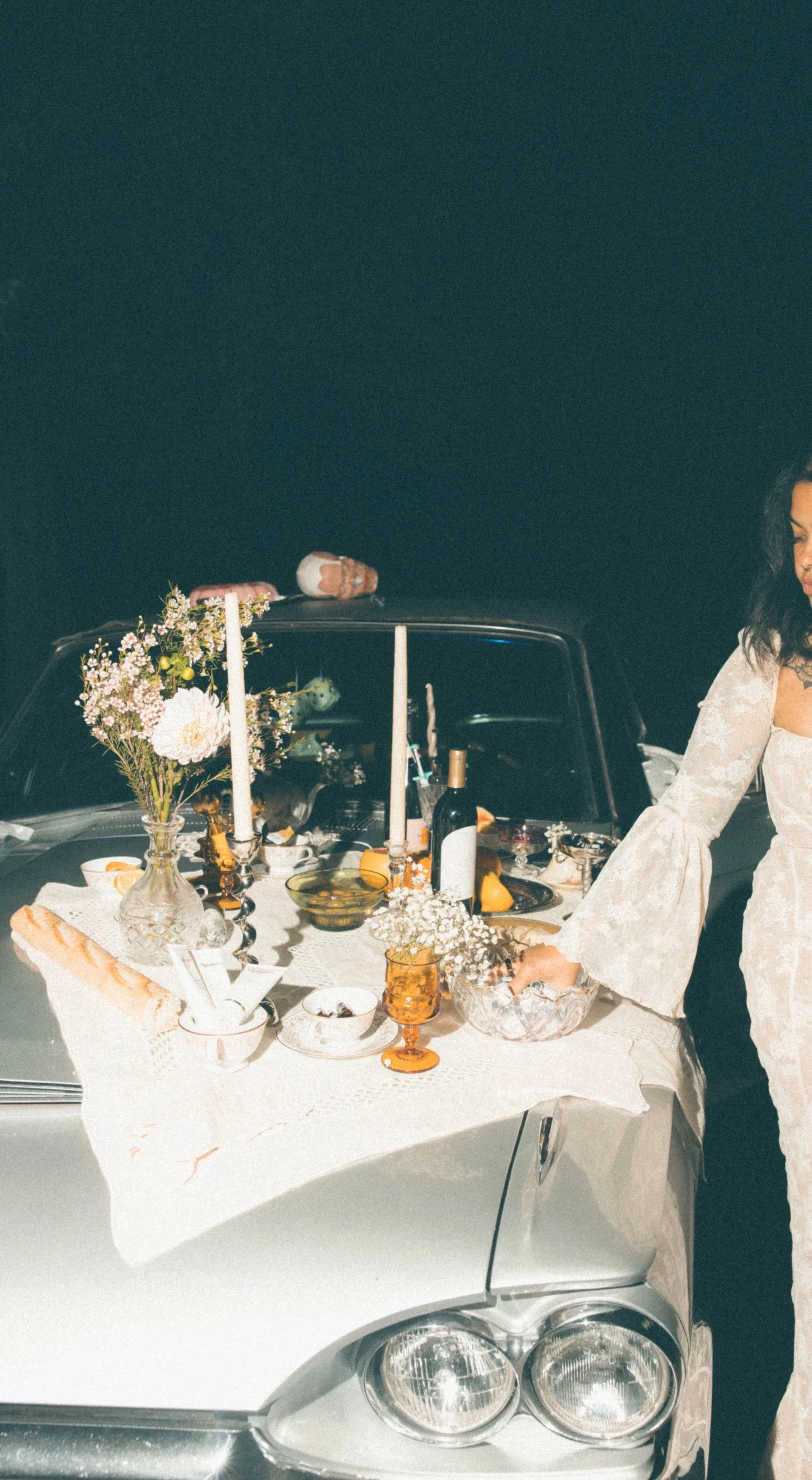 Table set with flowers, candles, wine, food, and dishes, arranged on the hood of a classic car with a woman in a cream lace dress.
