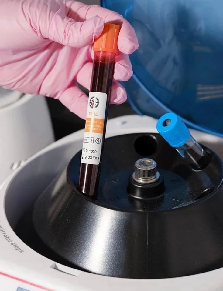 Person wearing pink gloves inserting a blood sample tube into a centrifuge machine while another unused tube with a blue cap is nearby.
