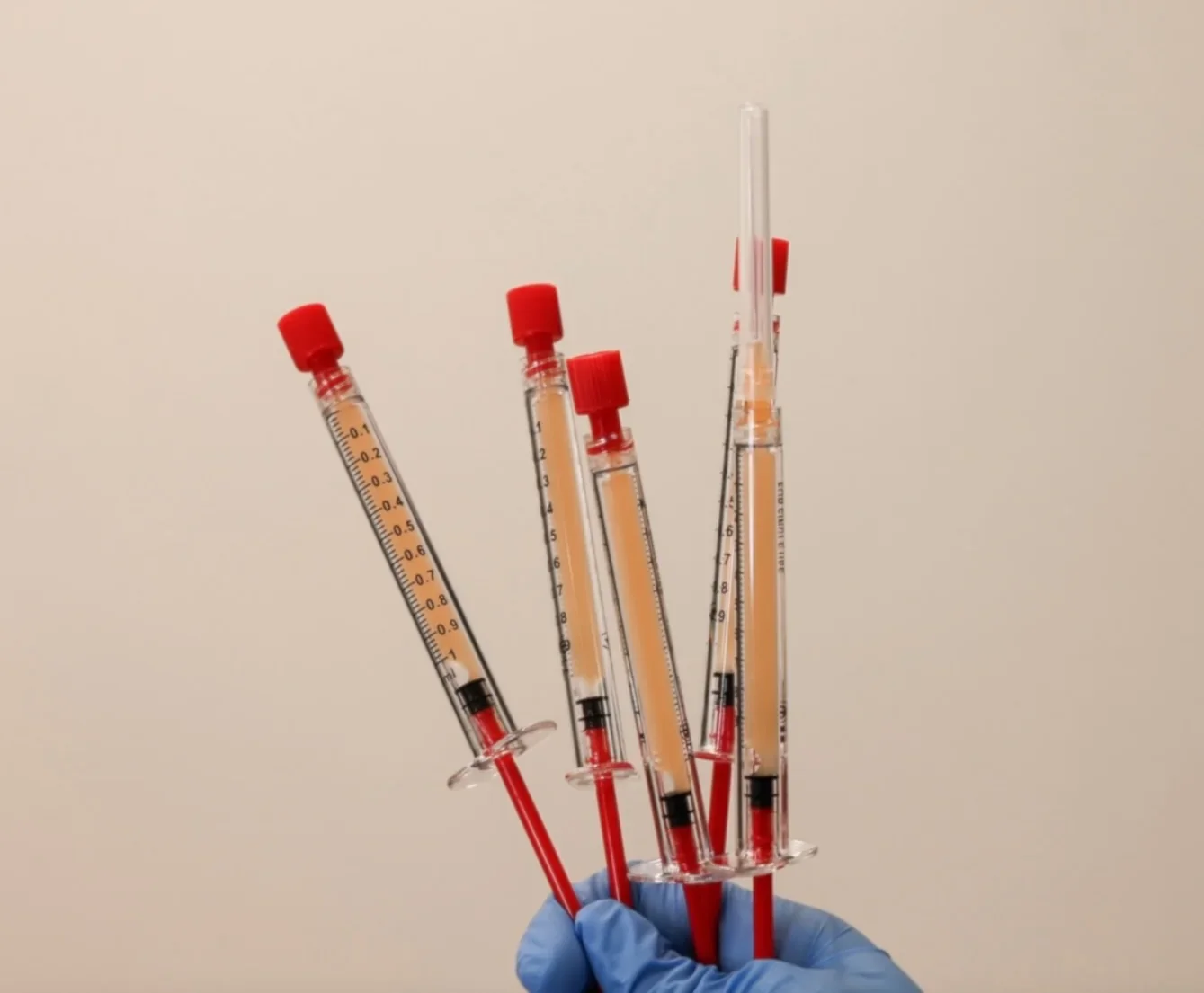 A gloved hand holding multiple syringes filled with orange-colored liquid.