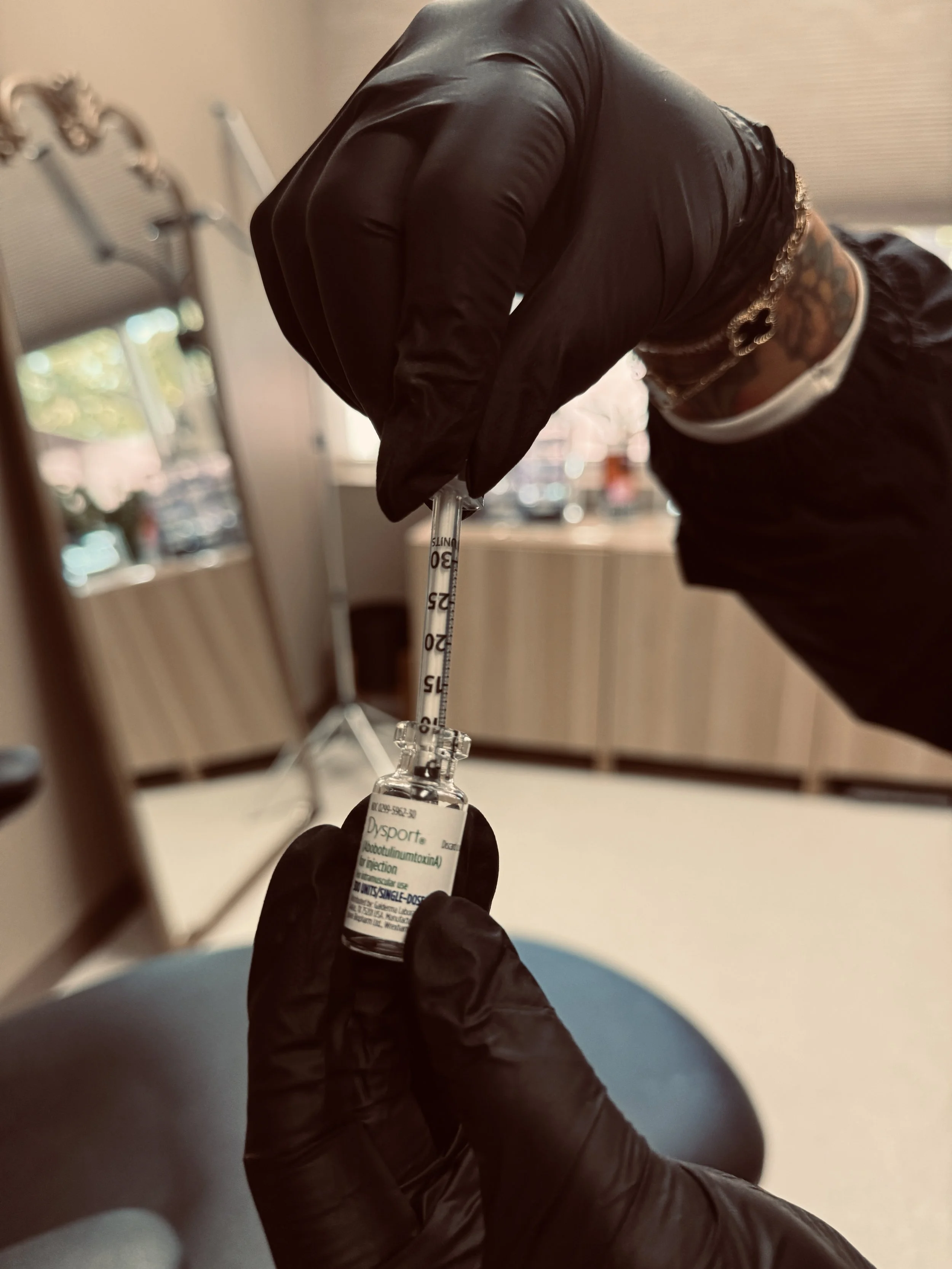 Person wearing black gloves filling a medical syringe with a vaccine or medication from a small vial in a clinical setting.