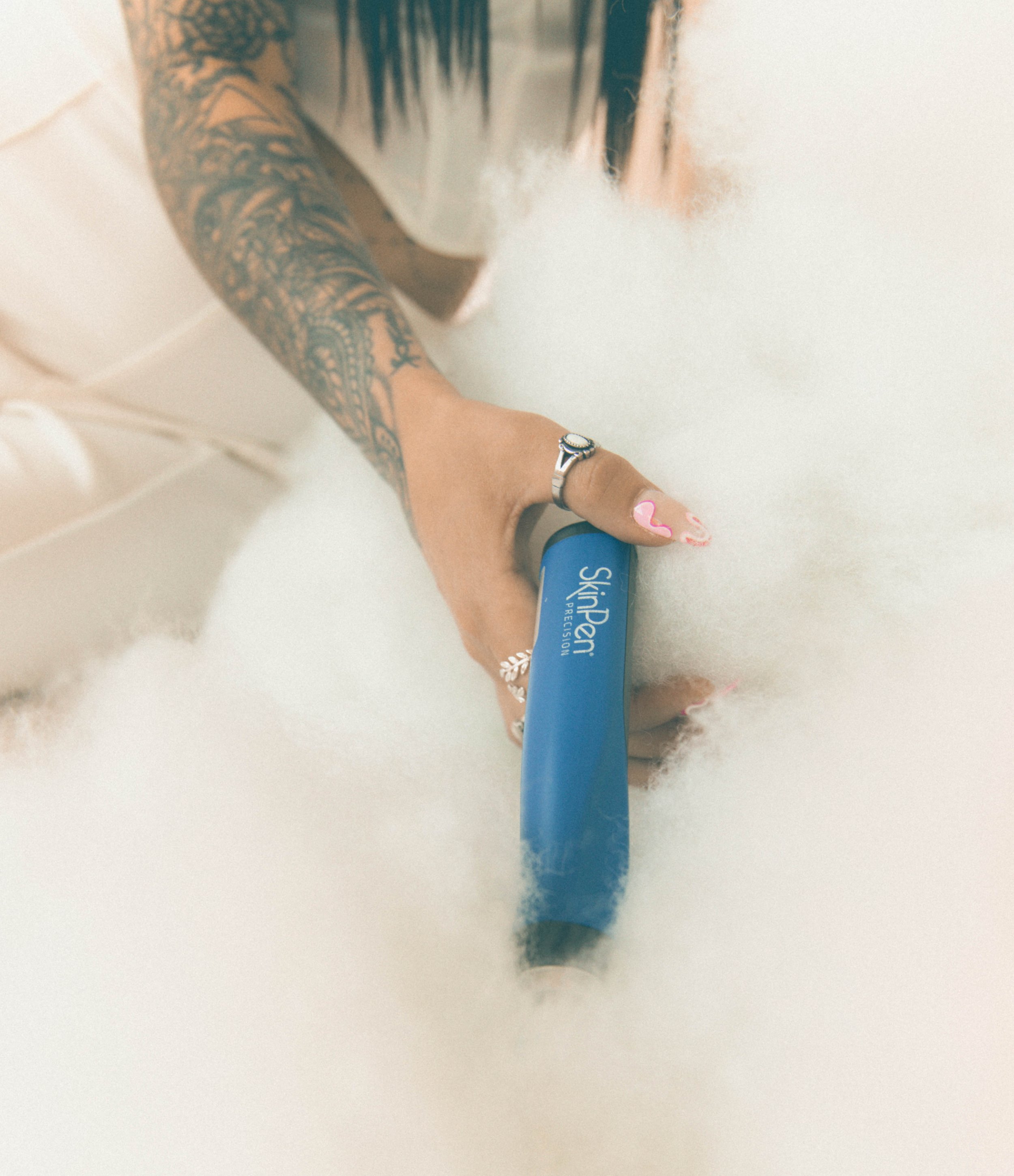 Person holding a Scotch-brite cleaning tool with their hand, surrounded by white foam or bubbles, tattooed arm, wearing rings, with painted nails.